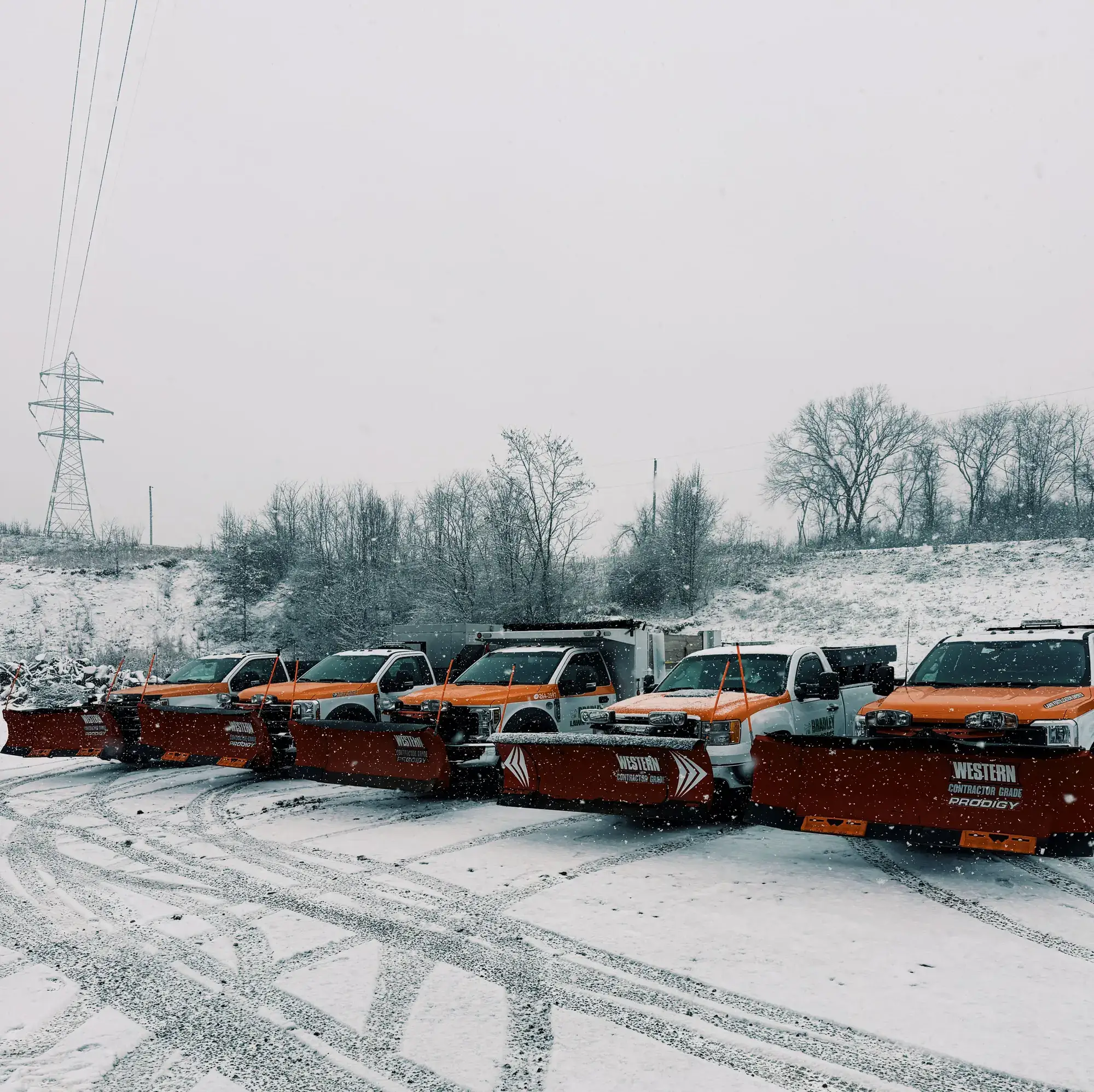 Row of plow trucks parked in the snow beside a utility corridor